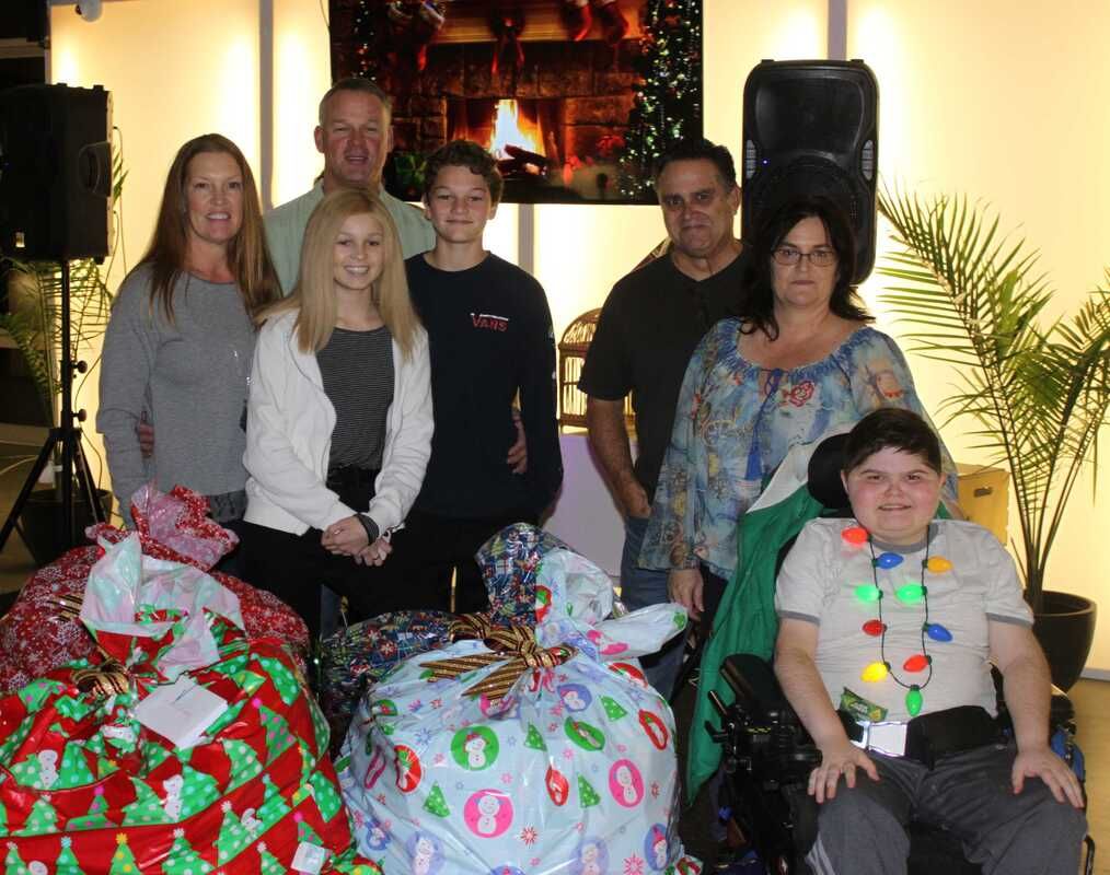 Group of people with wrapped gifts, posing near a speaker and a festive backdrop. One person uses a wheelchair.