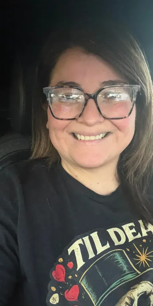 A person smiling while sitting in a car, wearing glasses and a dark graphic sweatshirt.