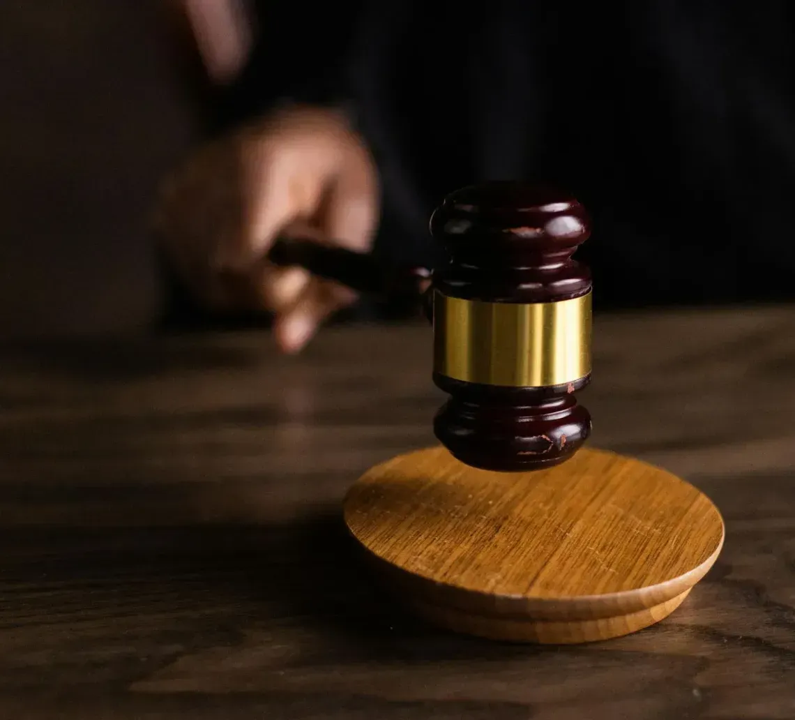 A wooden gavel with a gold band strikes a small wooden base in a courtroom setting.