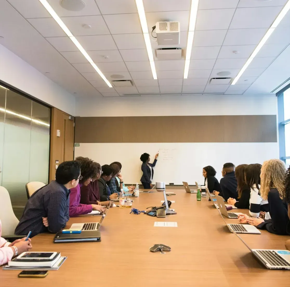 A diverse group of people attend a meeting around a long wooden conference table as one person presents at a whiteboard.