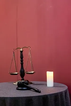 A pair of scales and a judge's gavel on a table next to a lit white candle against a deep red background.