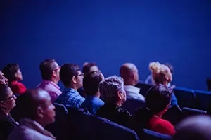 A diverse audience sits in a dimly lit auditorium, watching a presentation against a solid blue background.
