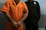 A person in an orange jumpsuit is handcuffed while being escorted by a uniformed law enforcement officer.