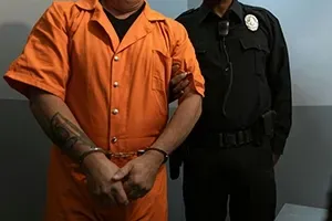 A person in an orange jumpsuit is handcuffed while being escorted by a uniformed law enforcement officer.