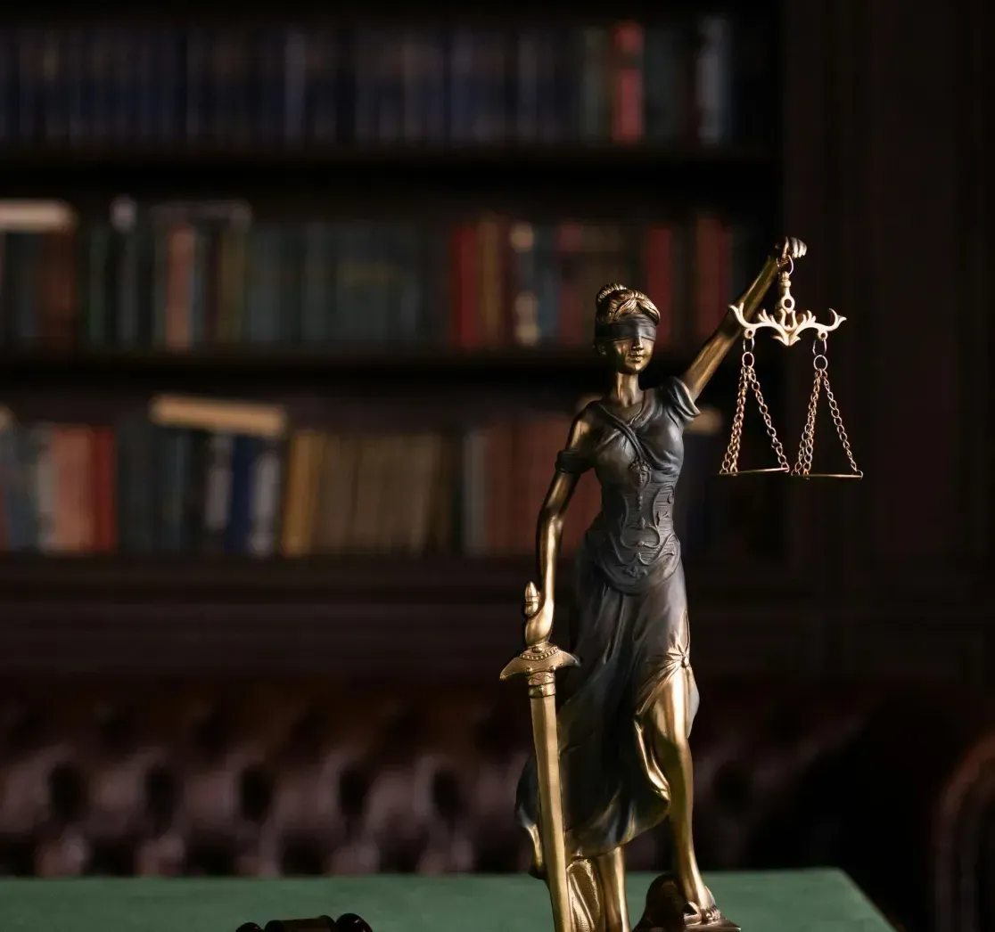 A statue of Lady Justice with a sword and scales stands on a green table next to a wooden gavel in a library.