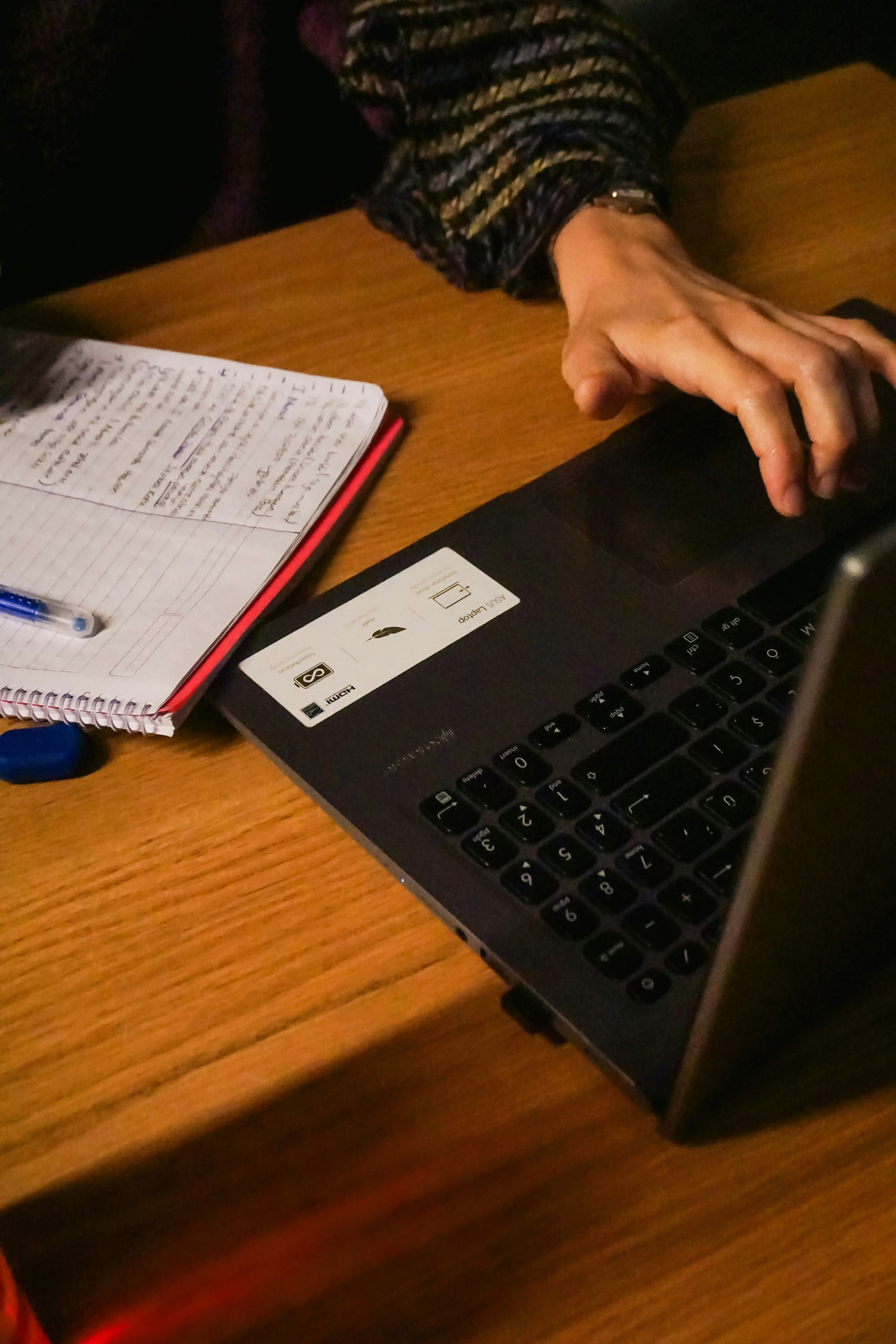 A person’s hand rests on a laptop keyboard near a notebook and a pen on a wooden desk.