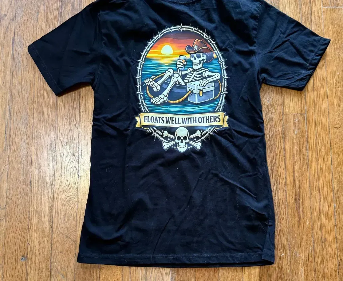 Black t-shirt featuring a graphic of a skeleton wearing a cowboy hat and floating on the water at sunset.
