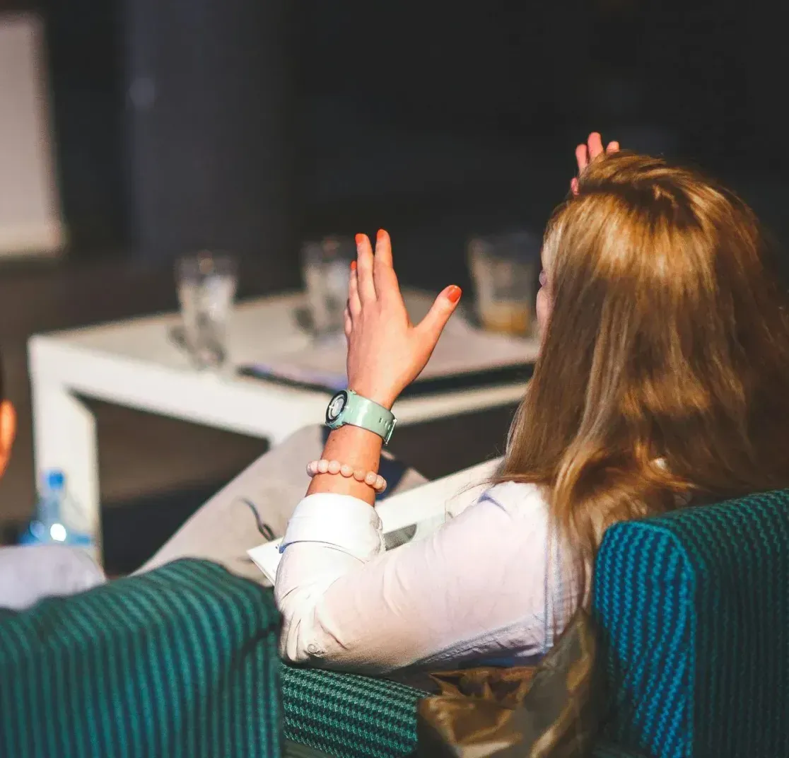 Two people in green chairs view a table with glasses during an indoor presentation. One person gestures while speaking.