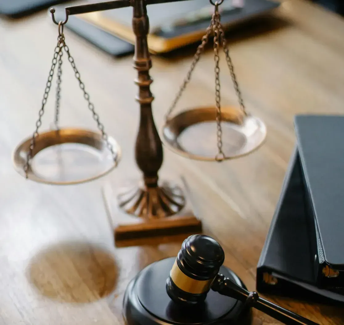A brass scale of justice and a wooden judge’s gavel resting on a wooden desk next to black legal binders.