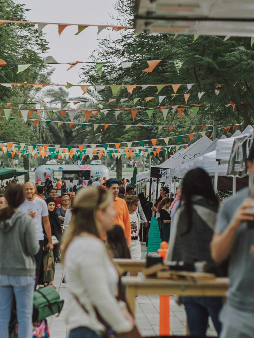 Crowd of people walking through an outdoor market decorated with rows of hanging green and orange triangular bunting.