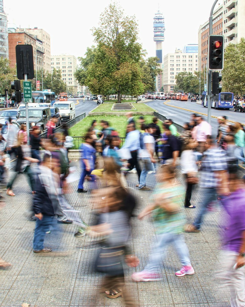 Pedestrians walk across a busy city street in front of a green median and a tall tower in the background.
