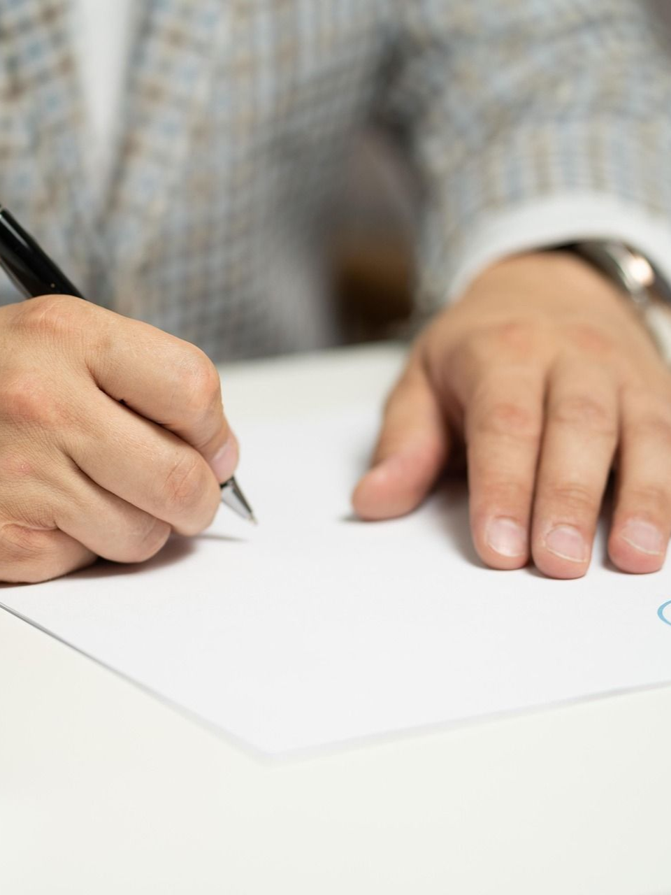 A person in a patterned blazer uses a pen to write on a blank white sheet of paper on a desk.