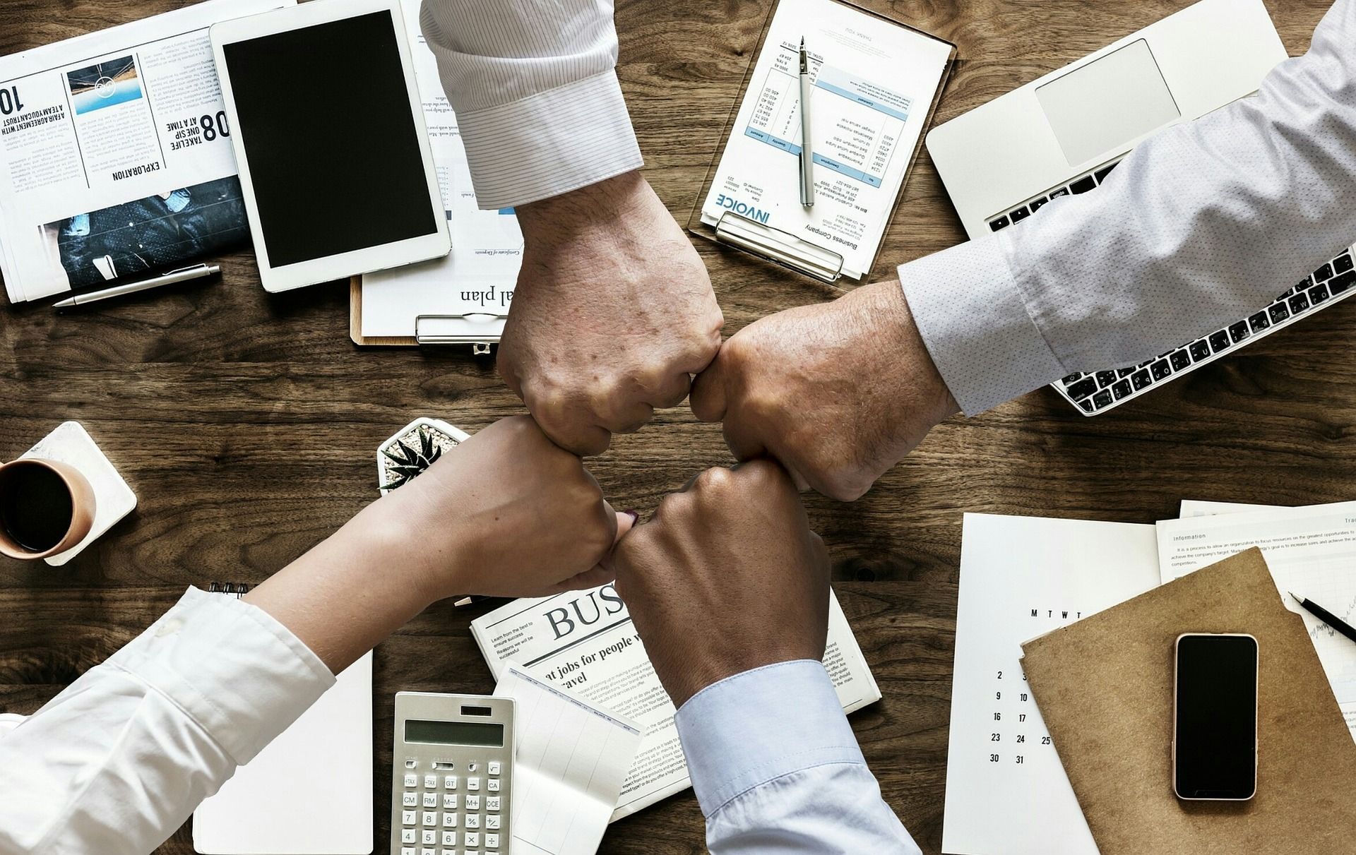 Four people in business attire join fists together in the center of a wooden desk scattered with papers and devices.