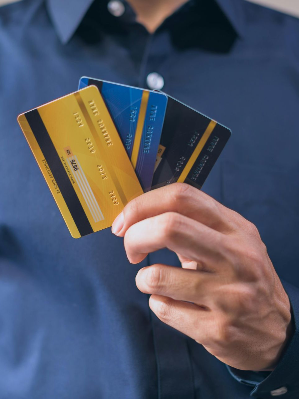A person in a blue shirt holds up three credit cards, showing their gold, blue, and black backs.
