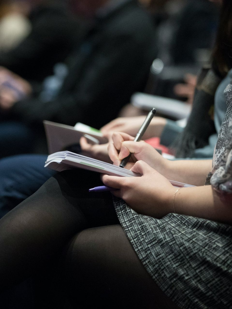 A person sits in a dim room, taking notes in a spiral-bound notebook with a pen.