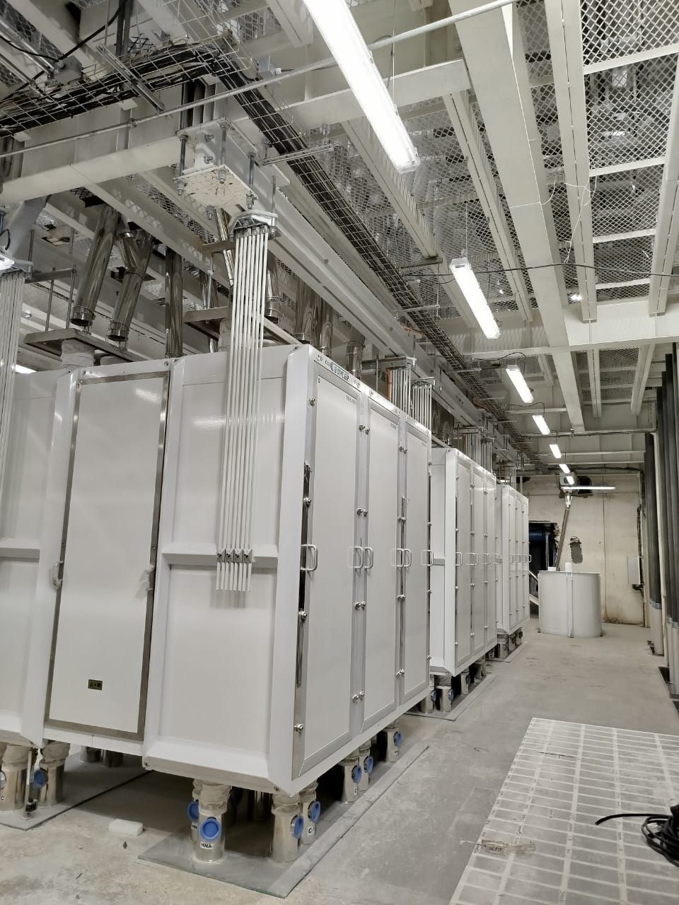 Fila de grandes máquinas industriales blancas en una instalación interior luminosa. Conductos y cables aéreos visibles.