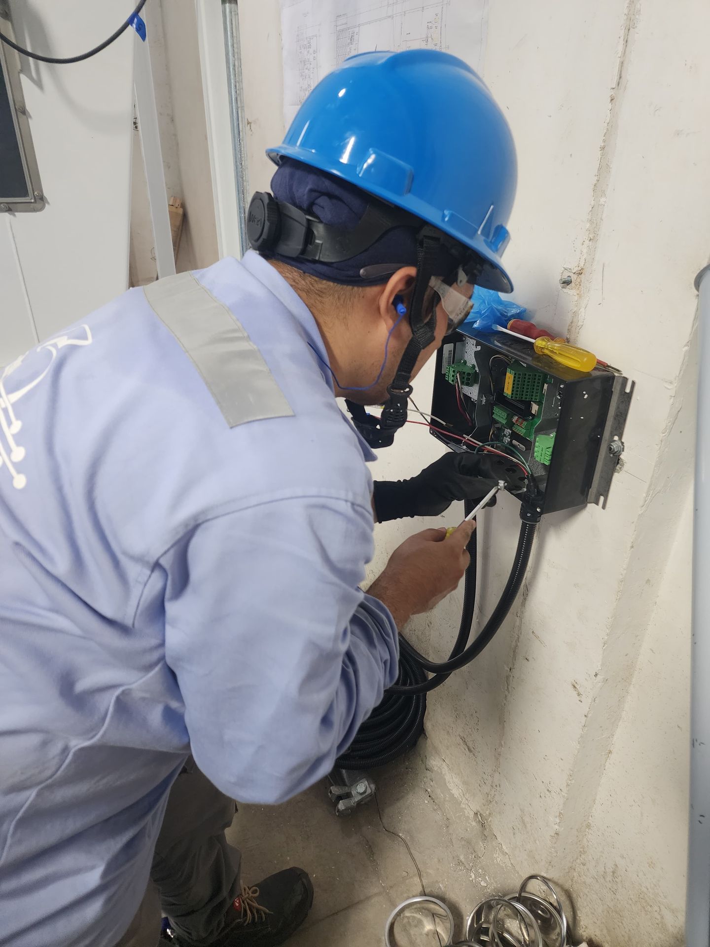 Electricista con casco azul trabajando en un panel montado en una pared de hormigón.