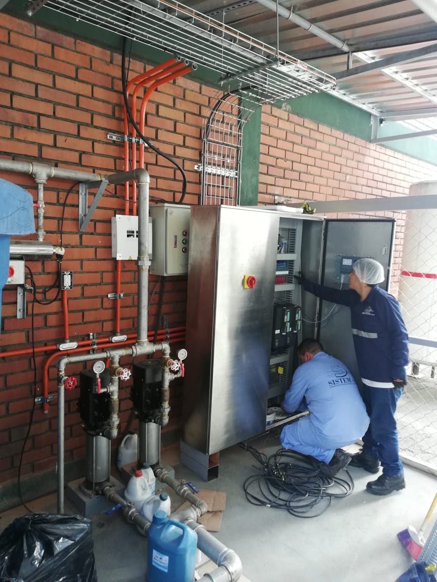Dos trabajadores con uniformes azules reparan un panel eléctrico junto a una pared de ladrillos
