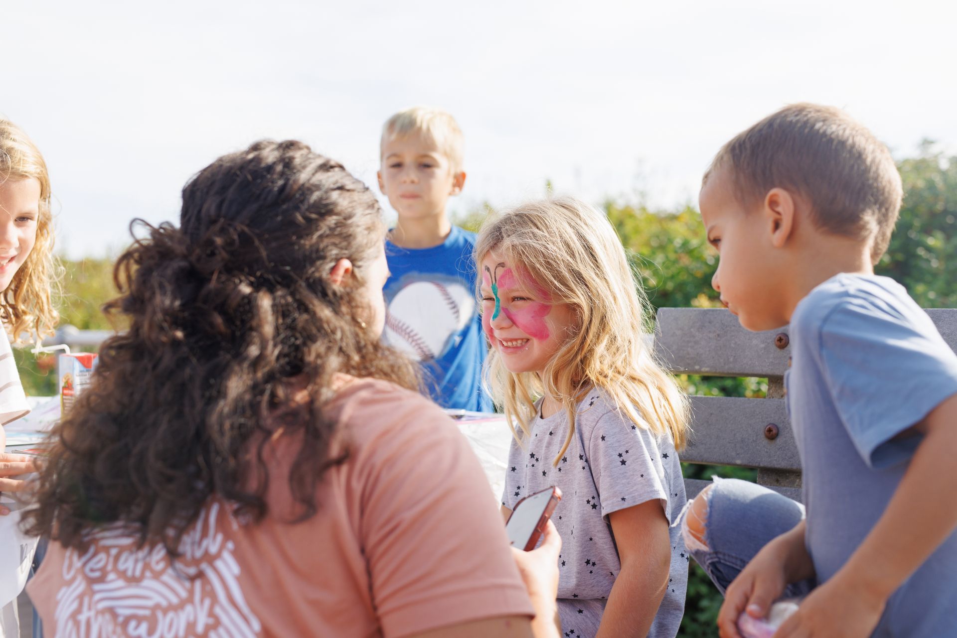 A person paints a girl's face at an outdoor event. Children look on with joy, in the sun.