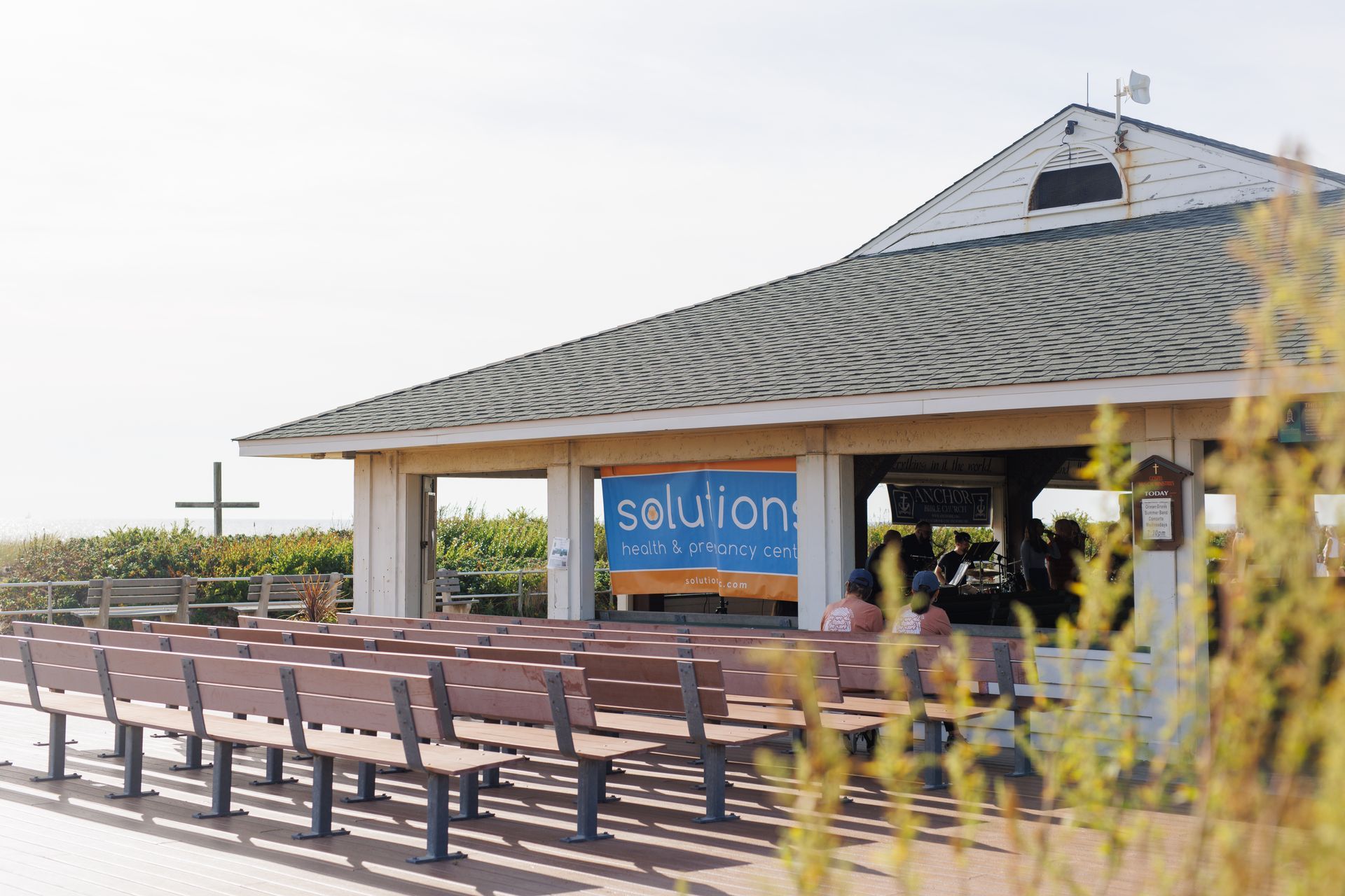 An outdoor pavilion with Solution banner, benches, and a cross in the background on a sunny day.