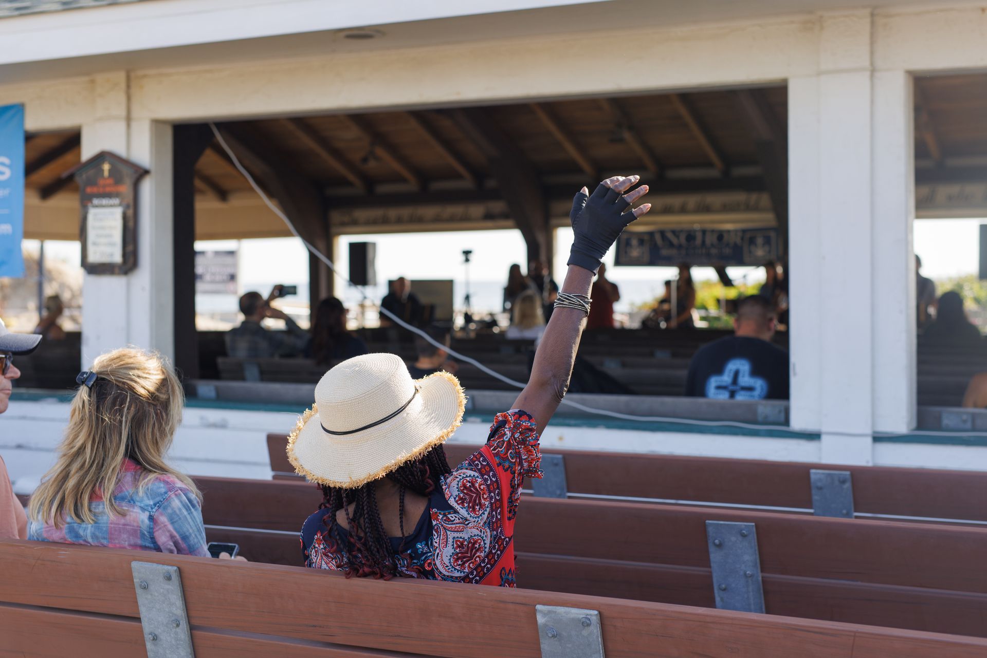 Person with straw hat and raised arm waves from bench at outdoor event.