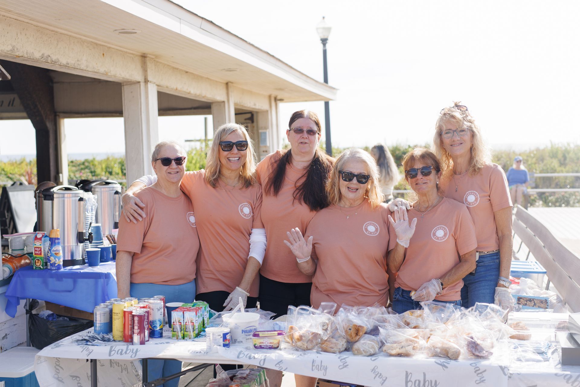 Group of seven women wearing matching orange shirts, at a table outdoors, smiling and waving.