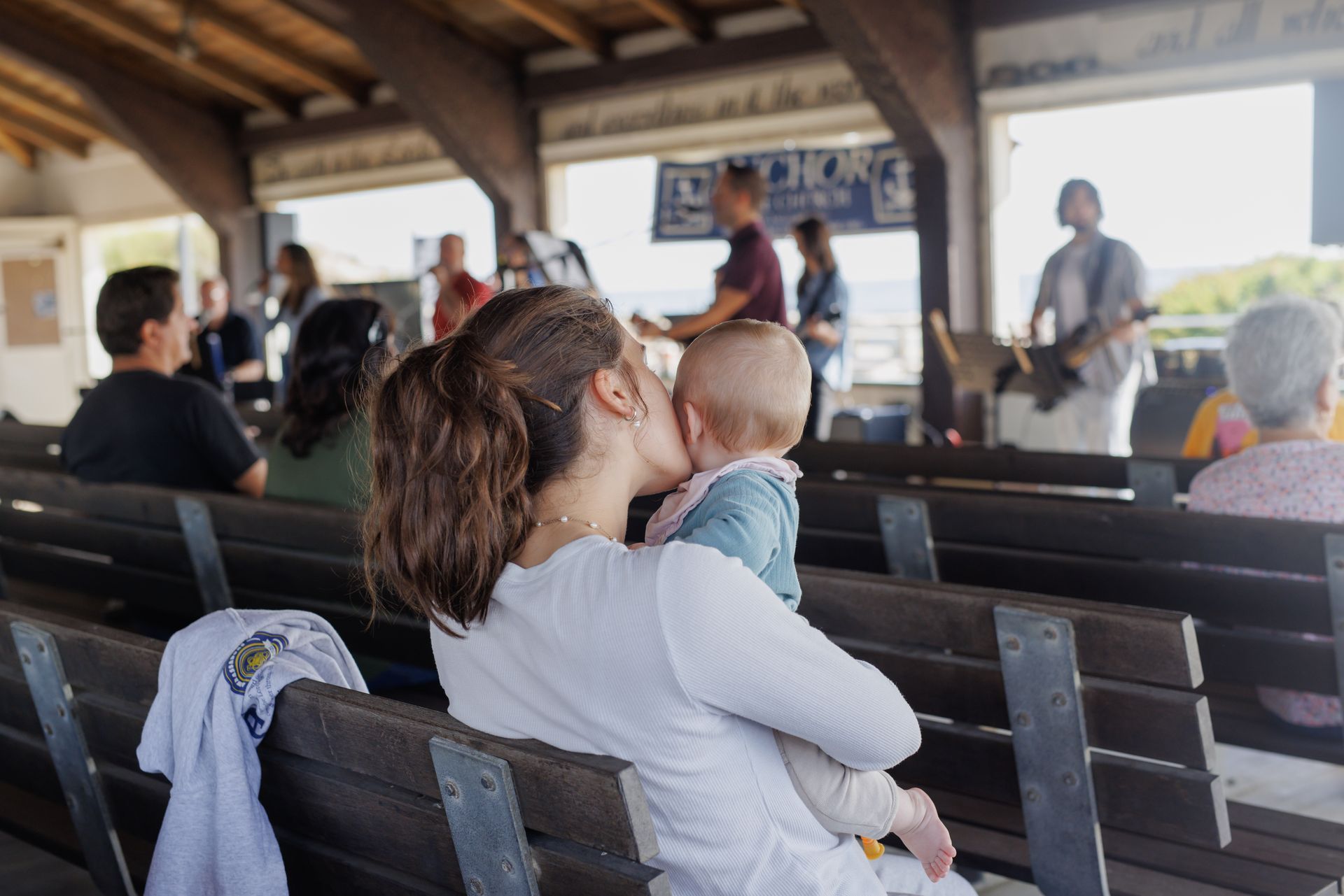 Woman kissing baby, watching band perform outdoors from wooden benches.