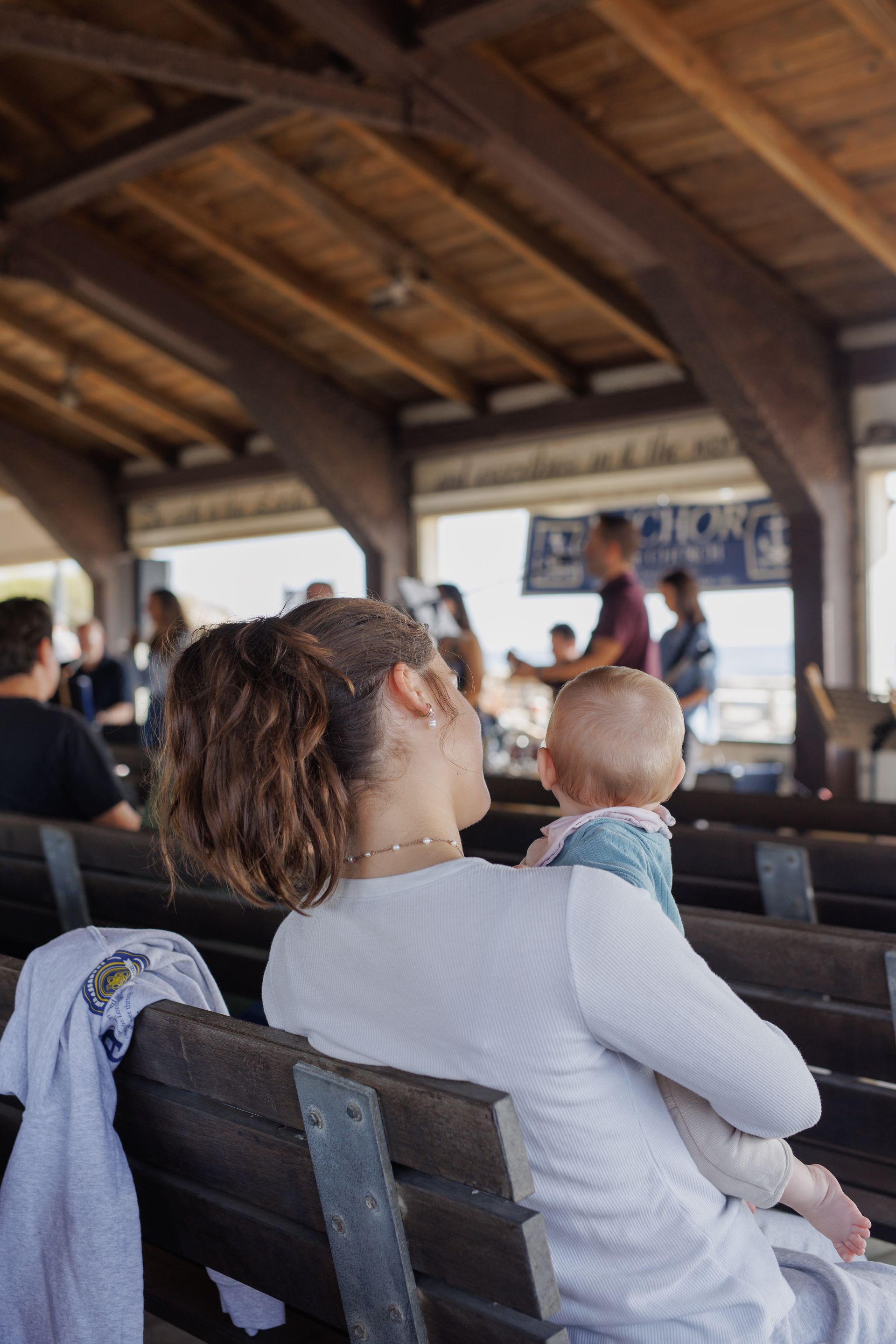 Woman holding a baby in an open-air building, looking toward other people, wood ceiling, and benches.