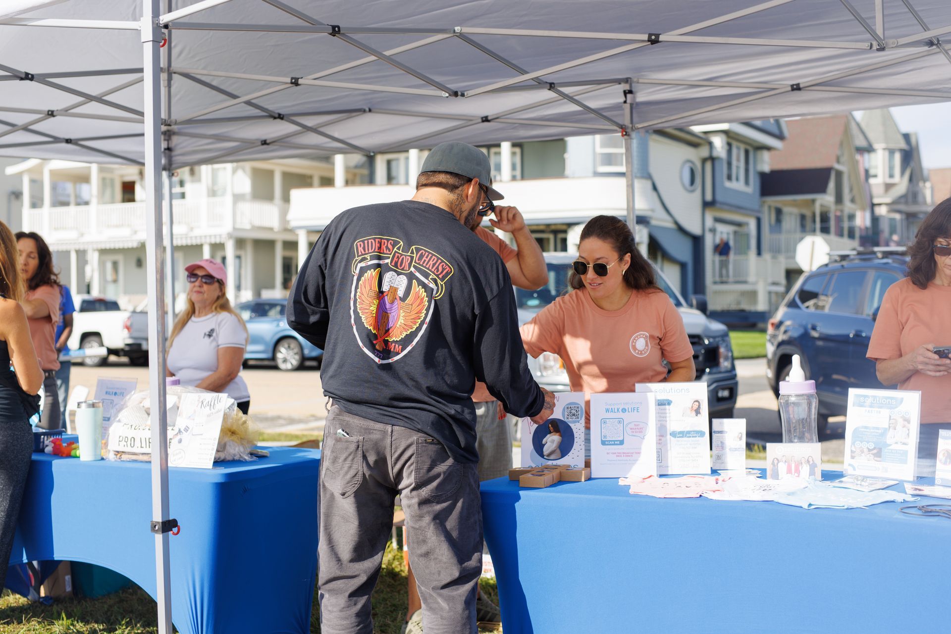 People at an outdoor booth with informational materials. Buildings and cars in the background.
