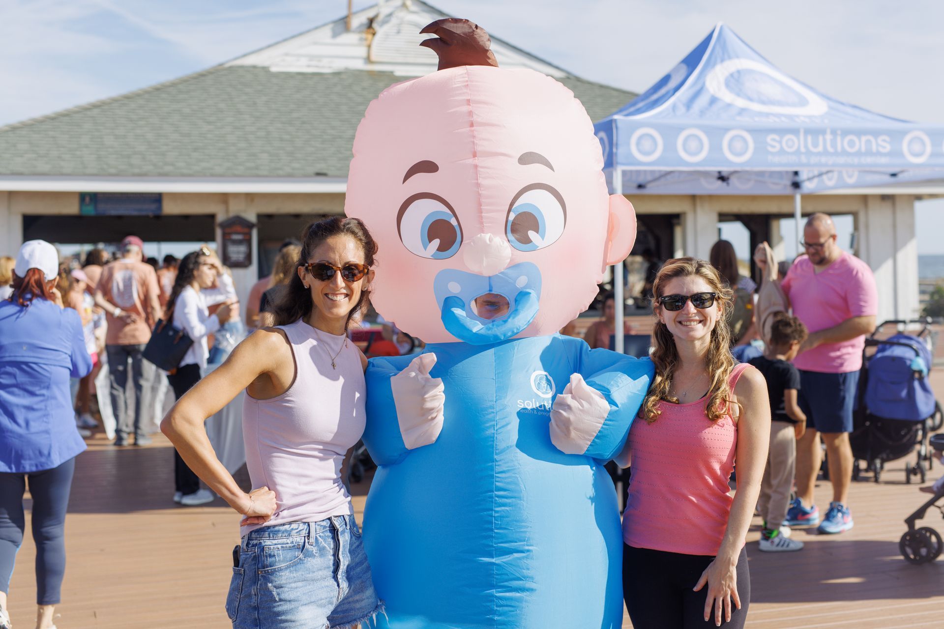 Two women pose with a blue baby mascot, smiling, at an outdoor event near a white building.