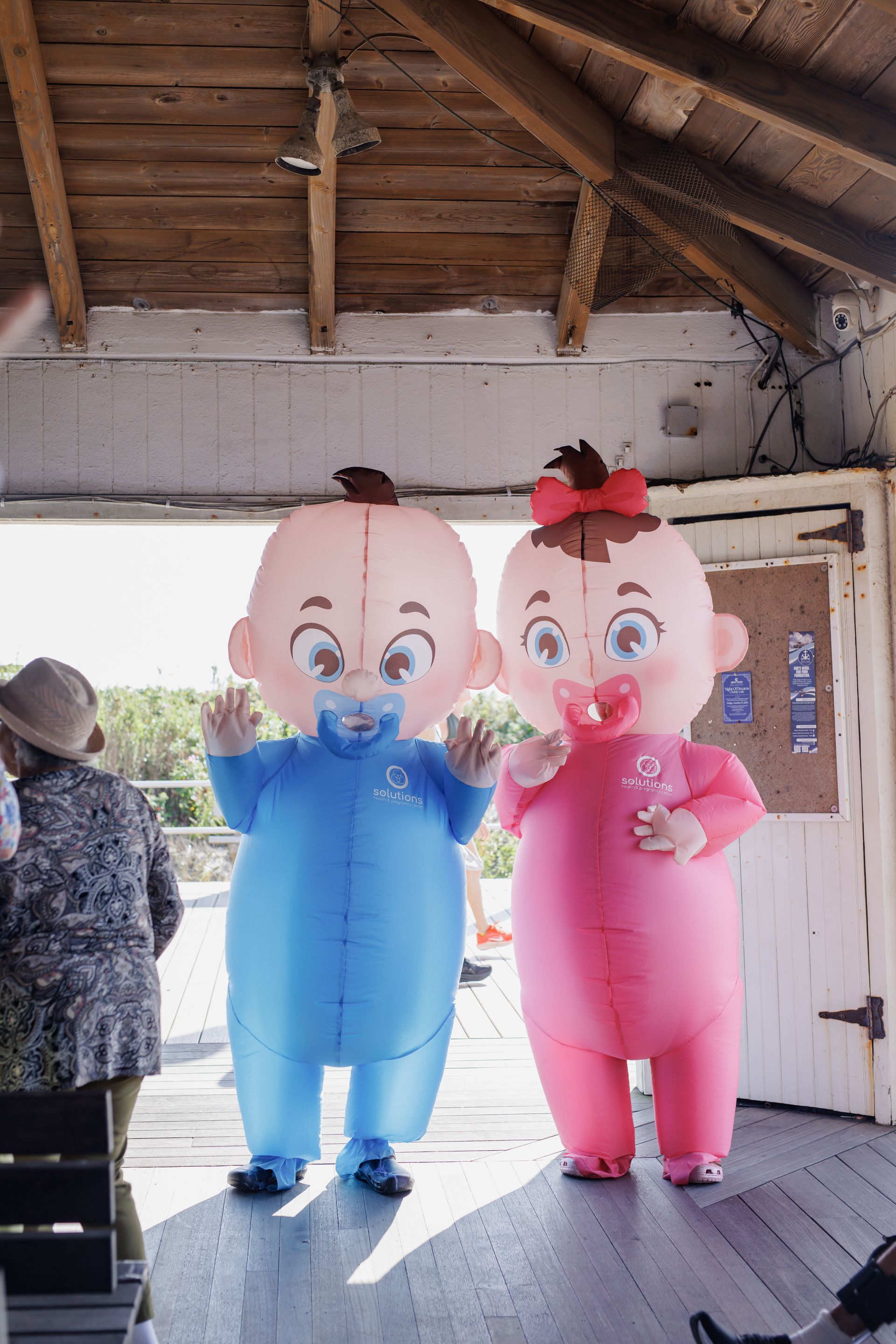 Two inflatable baby costumes, blue and pink, waving, in a wooden structure.