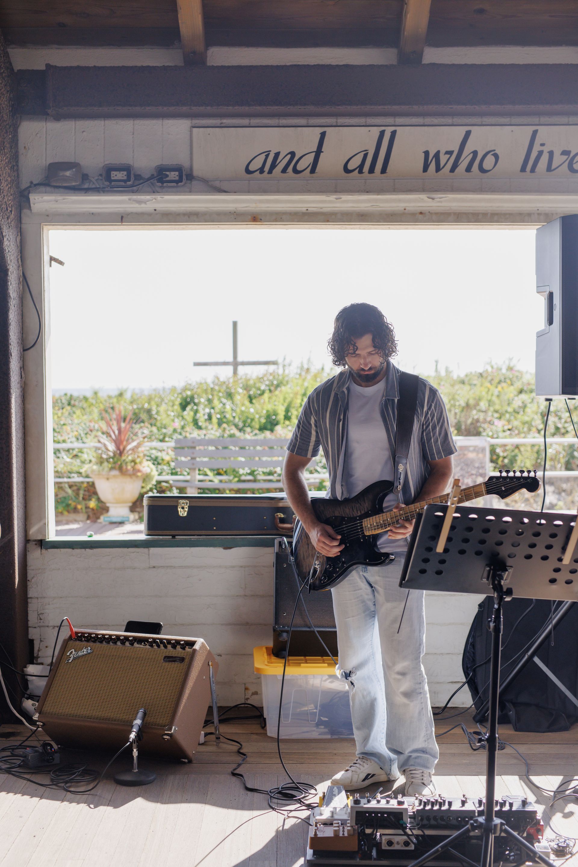 Man playing guitar outdoors. He is wearing a shirt, blue jeans, and white shoes.