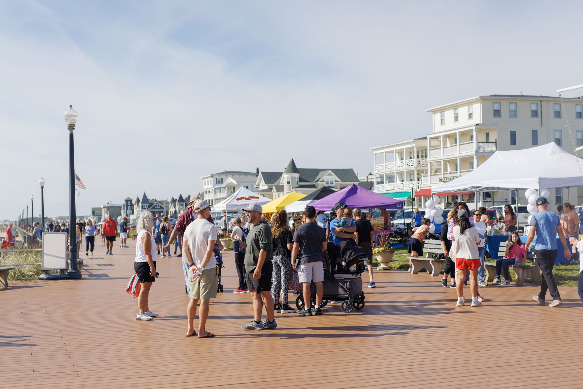 People gathered on boardwalk, tents, shops, and buildings in the background on a sunny day.