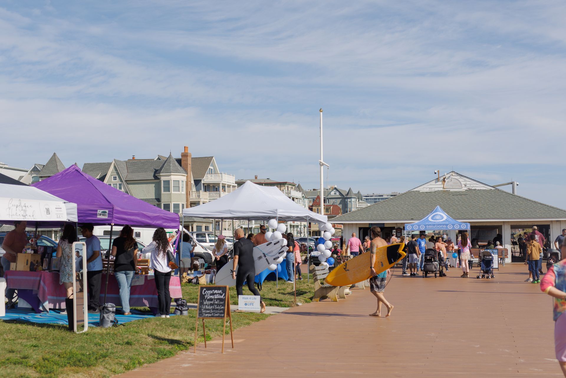 Outdoor fair with tents, people, and a person carrying a surfboard on a boardwalk near houses.