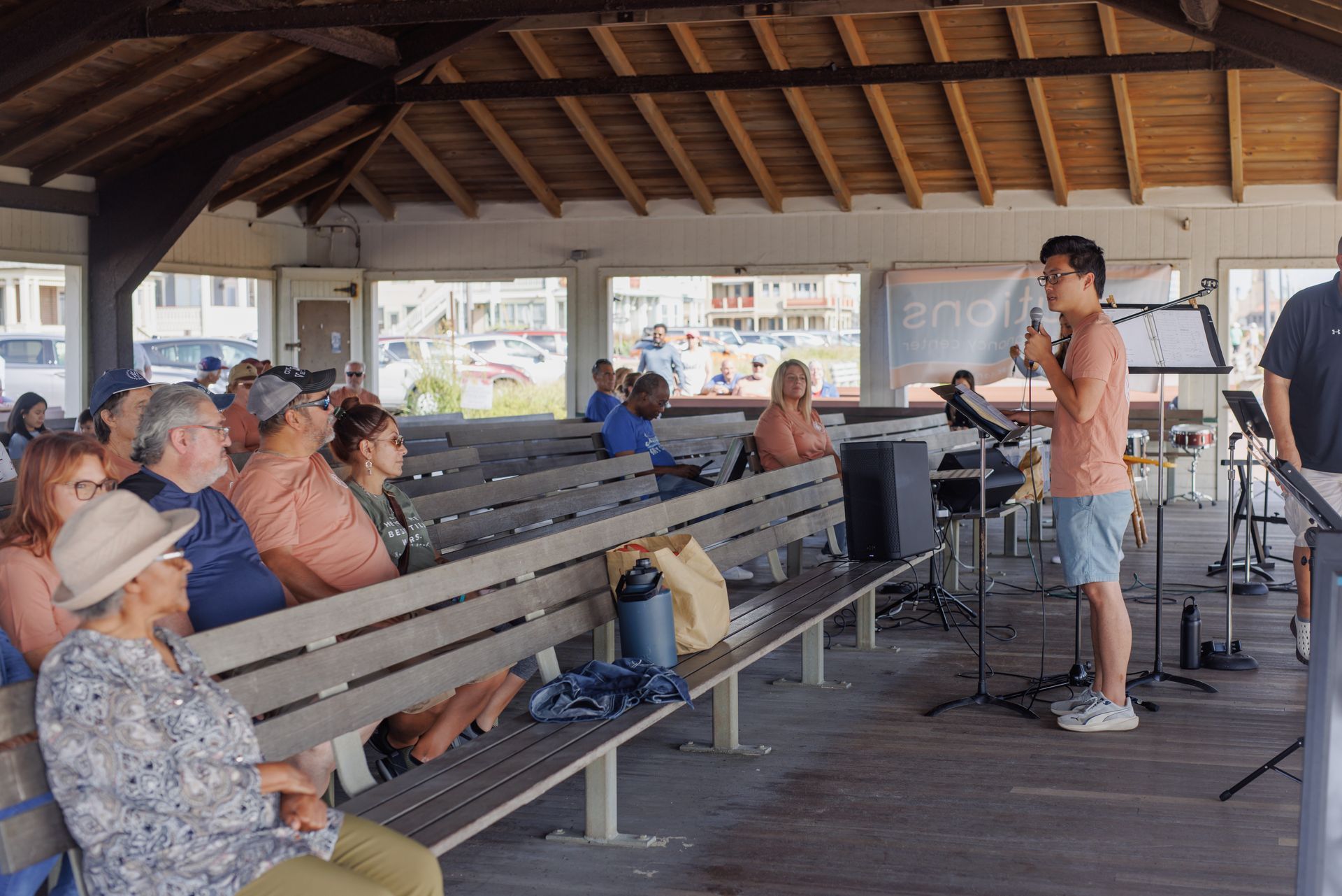 A man speaks into a microphone at an outdoor event with an audience seated on benches.