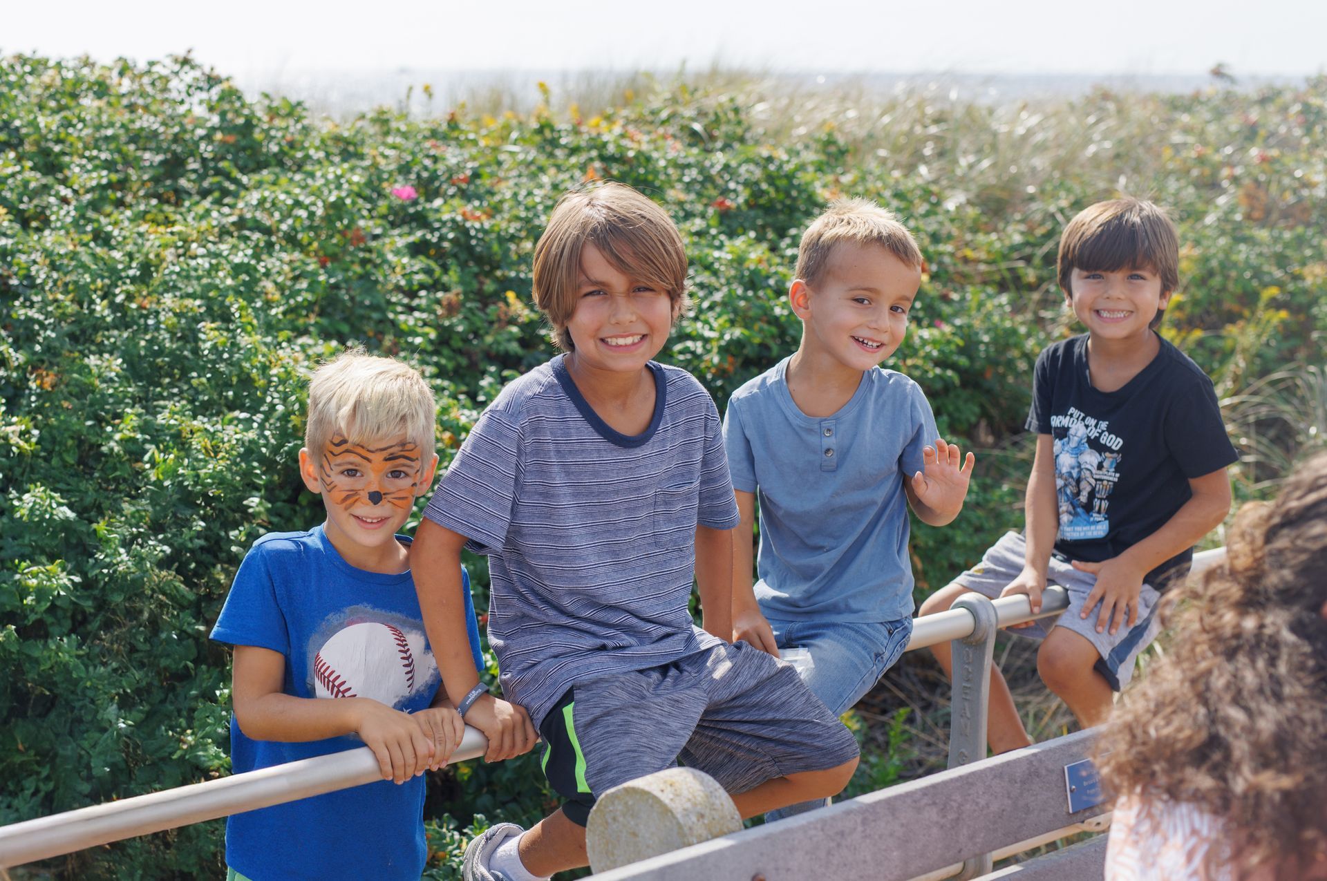 Four smiling boys pose on a wooden fence near the ocean, one with face paint.