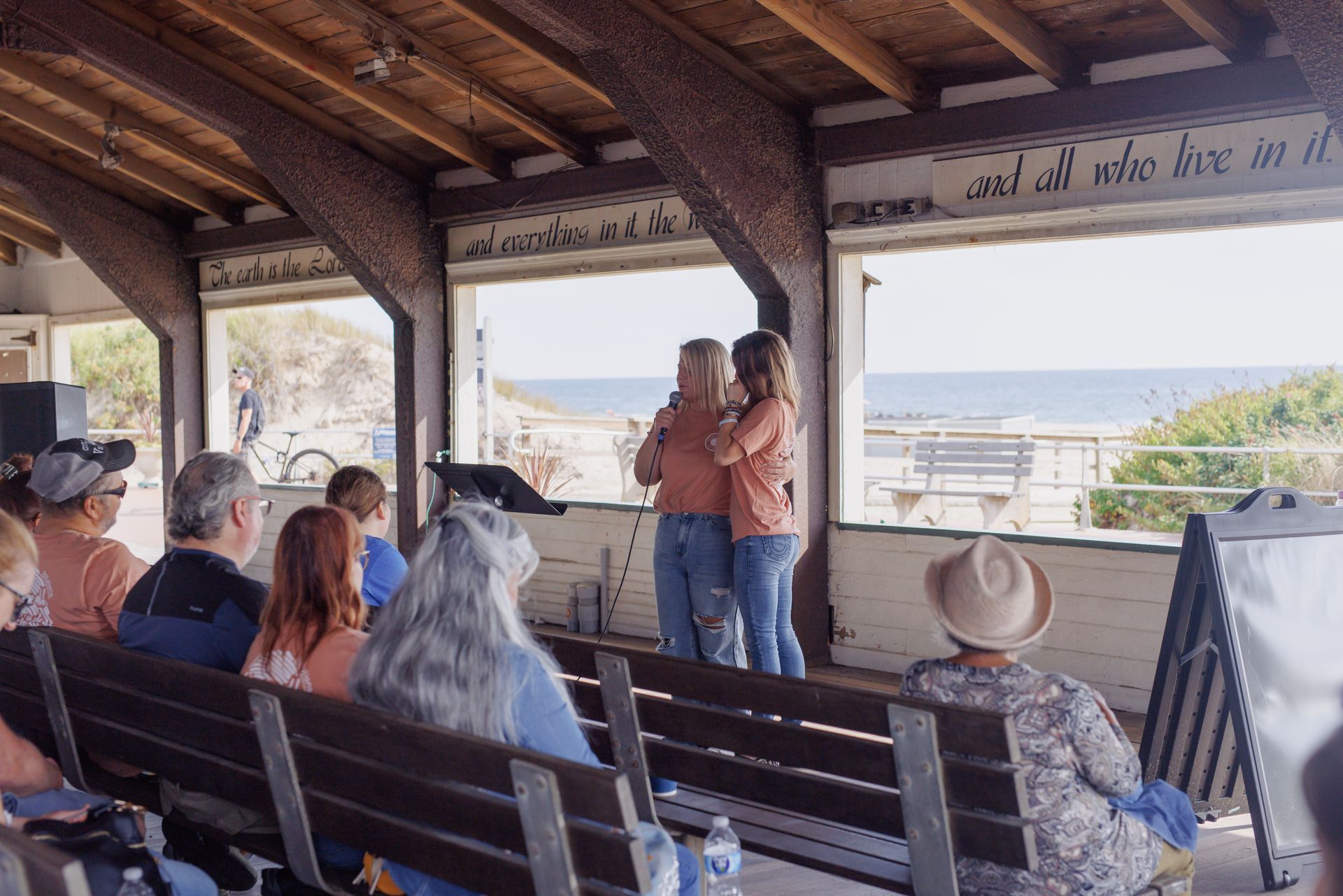 Couple hugging onstage by the ocean; audience watches from benches.