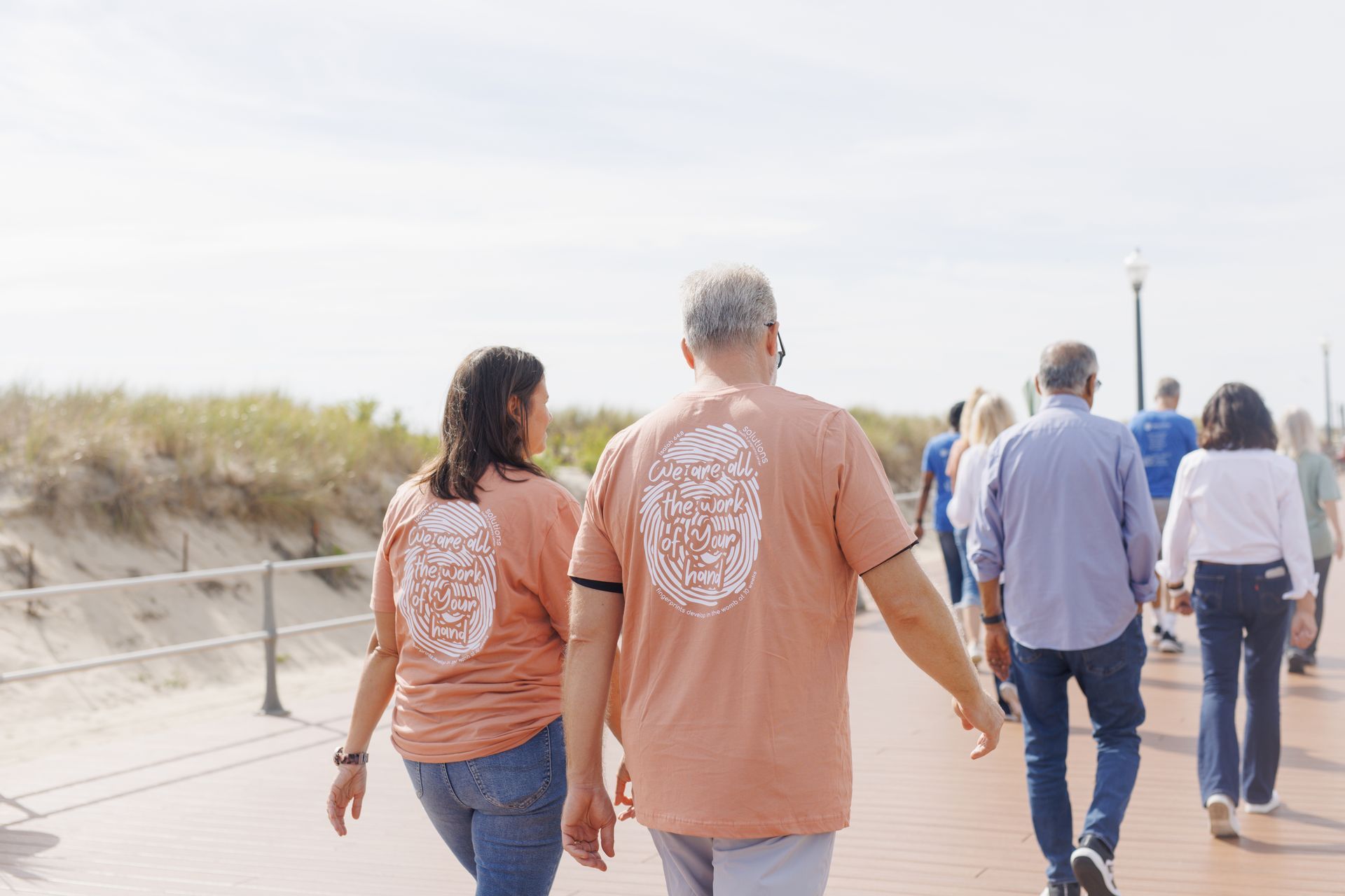 Two people walk on a boardwalk, wearing matching orange shirts with a design on the back, near the ocean.