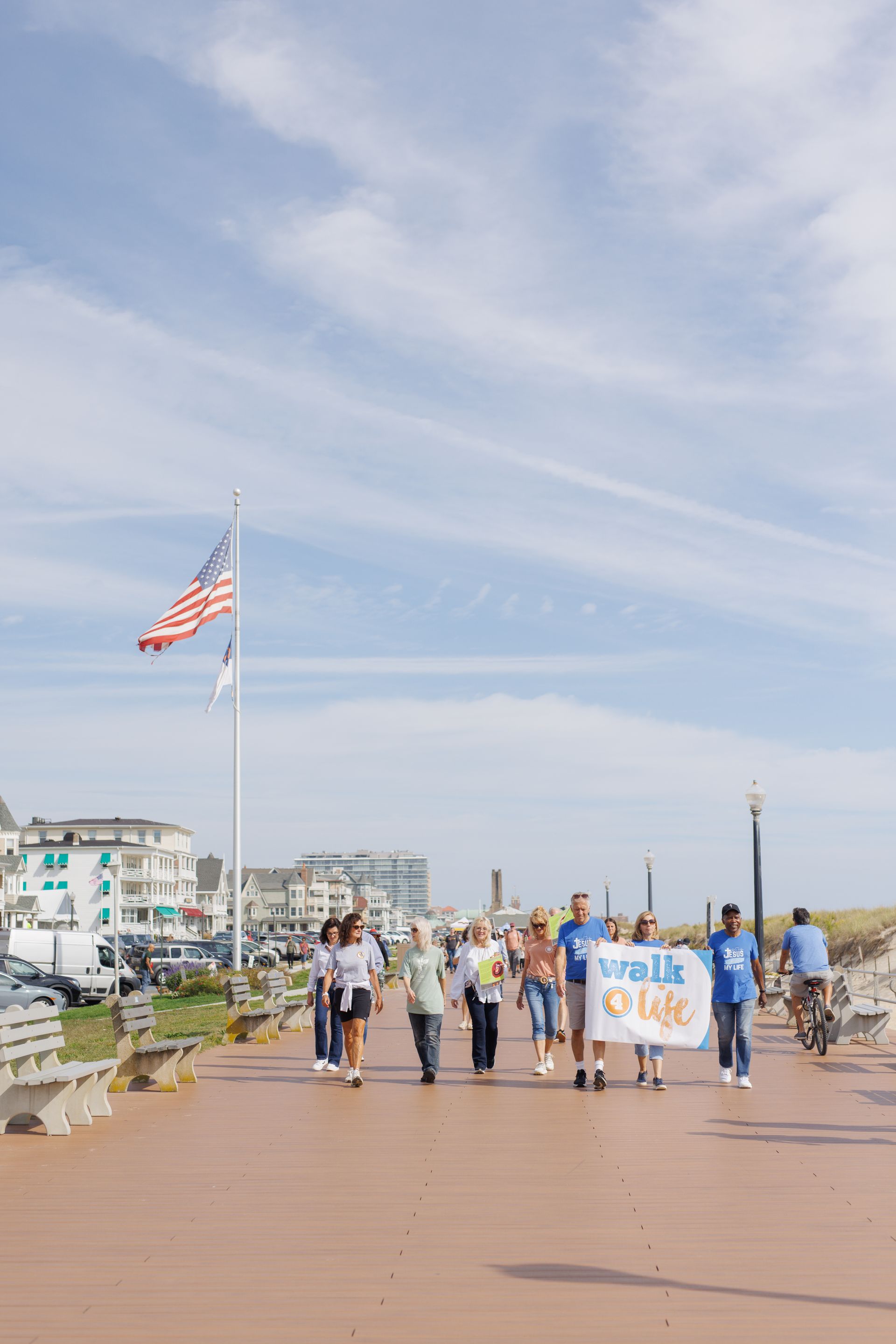 People walking on a boardwalk, carrying a banner, near a beach with a flag on a sunny day.
