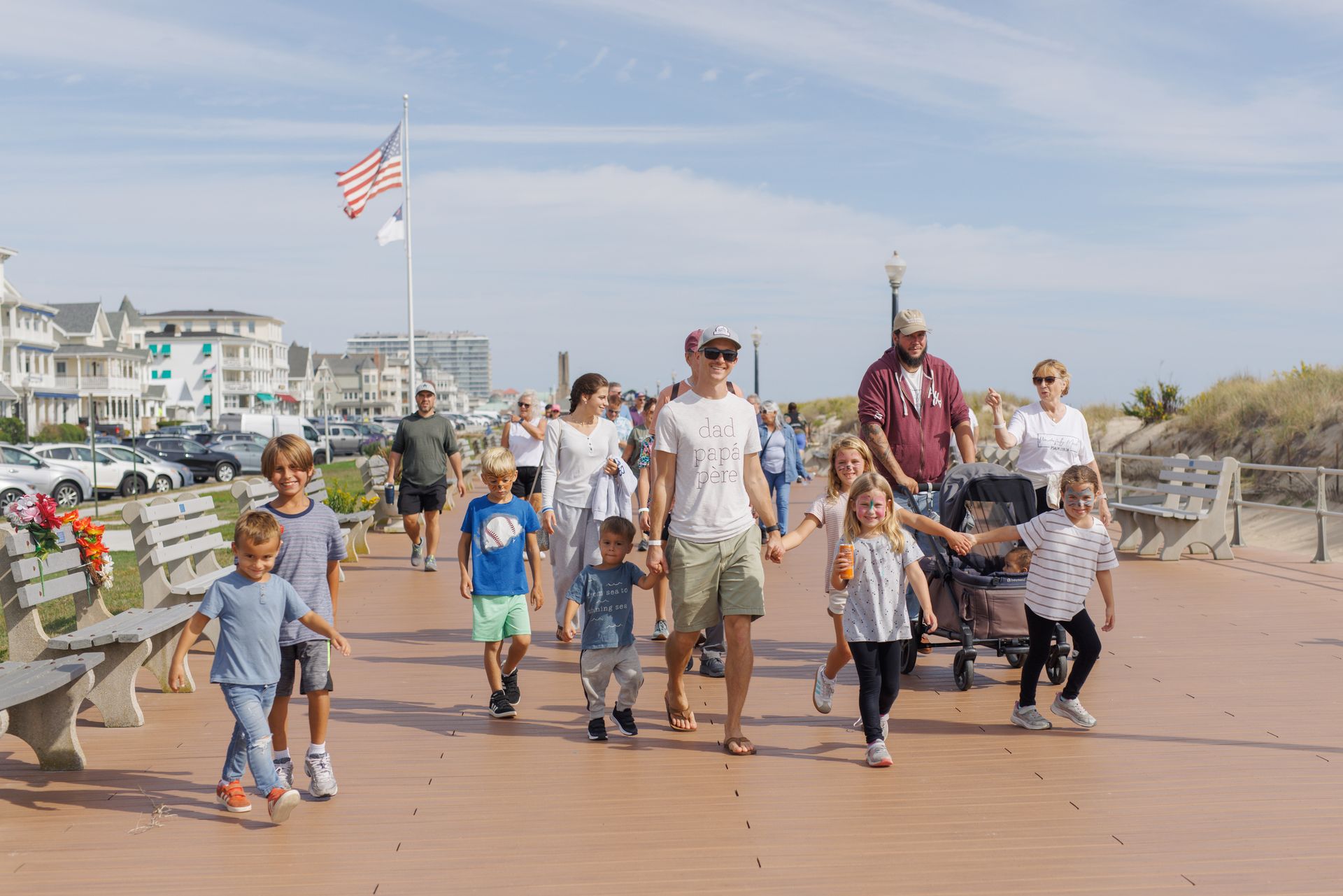 Family walking along a boardwalk, sunny day. Beach and buildings in the background. American flag flies overhead.