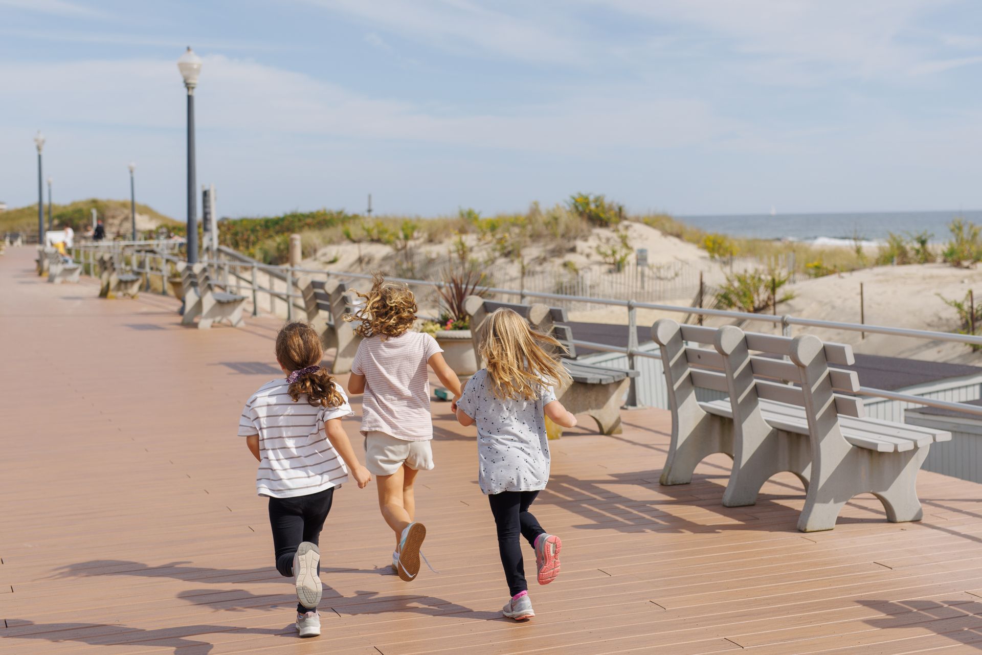 Three young girls running on a boardwalk towards the beach.