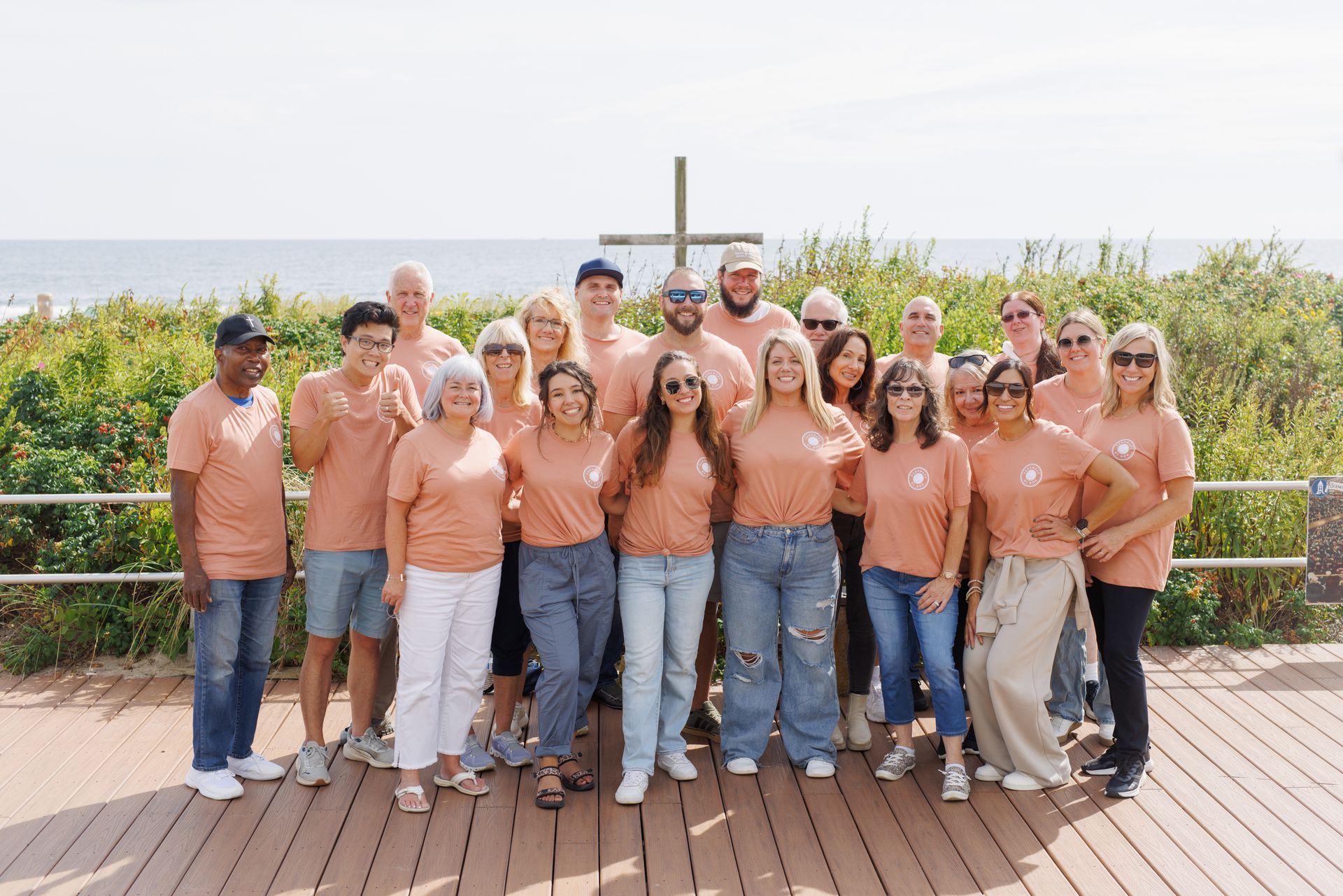 Group of people wearing peach shirts, standing in front of a wooden cross on a beach boardwalk.
