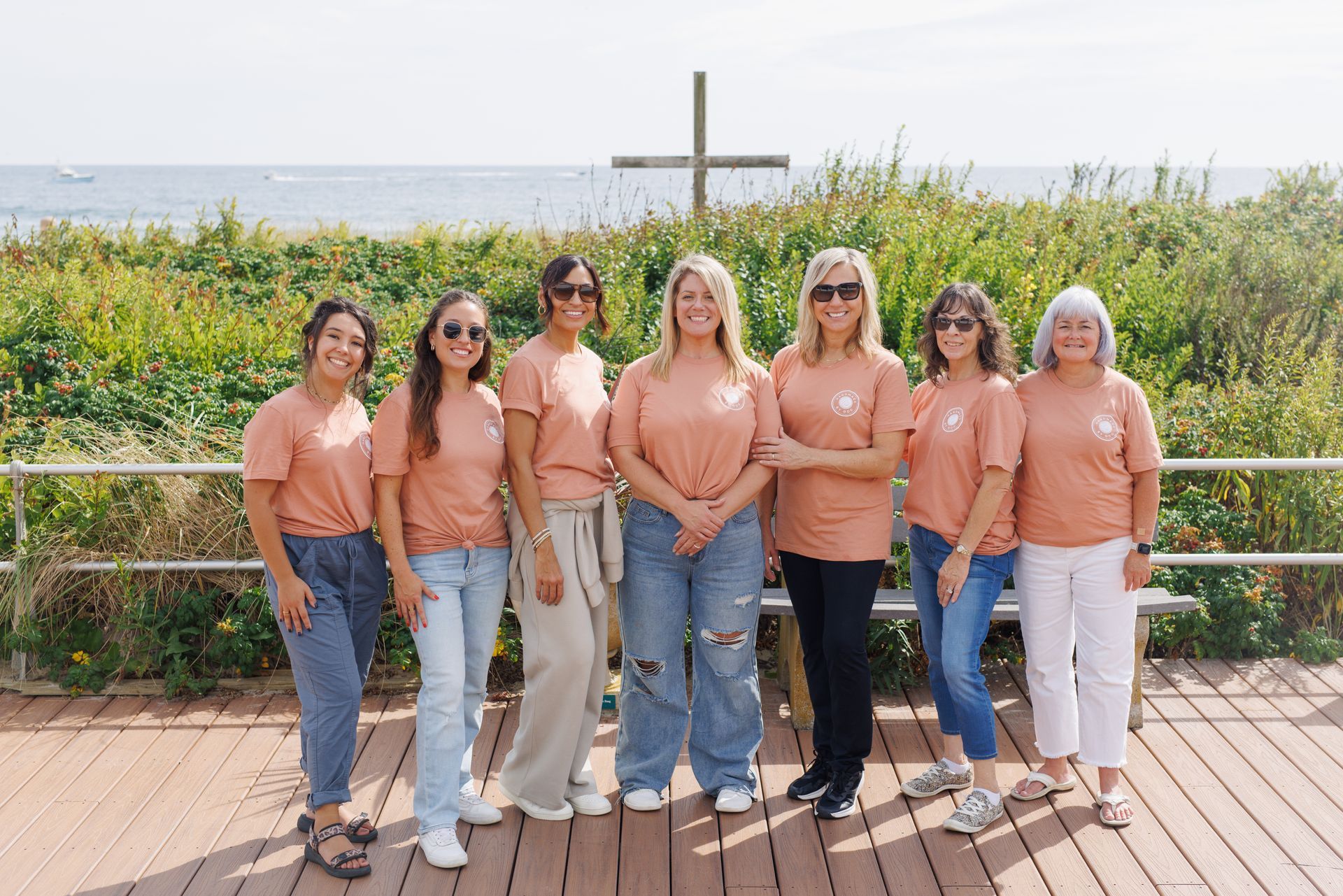 Seven women in matching peach shirts pose on a boardwalk with a cross and the ocean in the background.