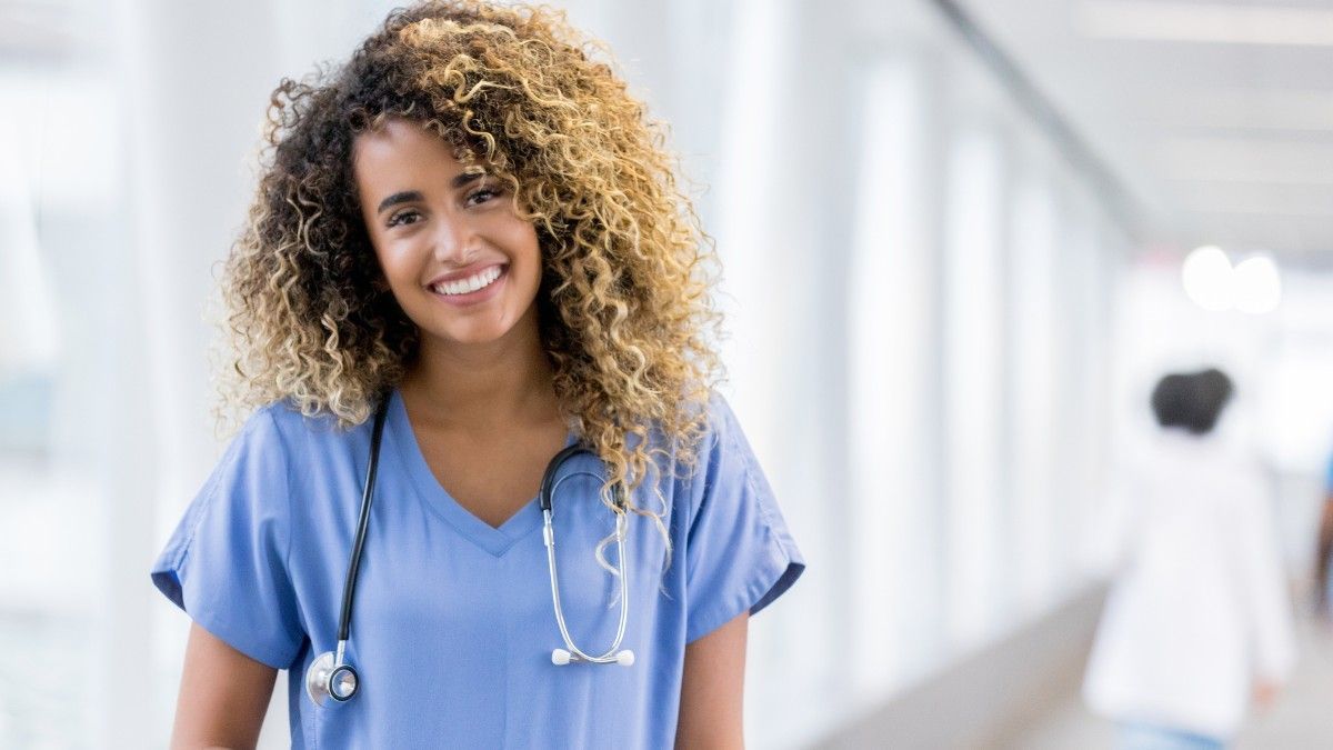 A nurse is standing in a hospital hallway with a stethoscope around her neck.