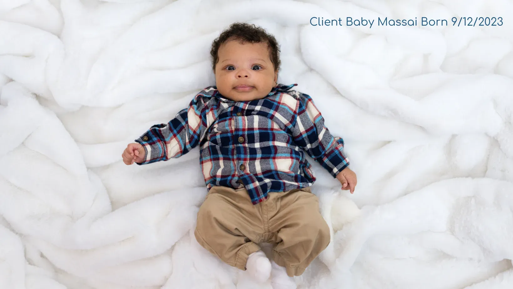 A baby in a plaid shirt is laying on a white blanket.
