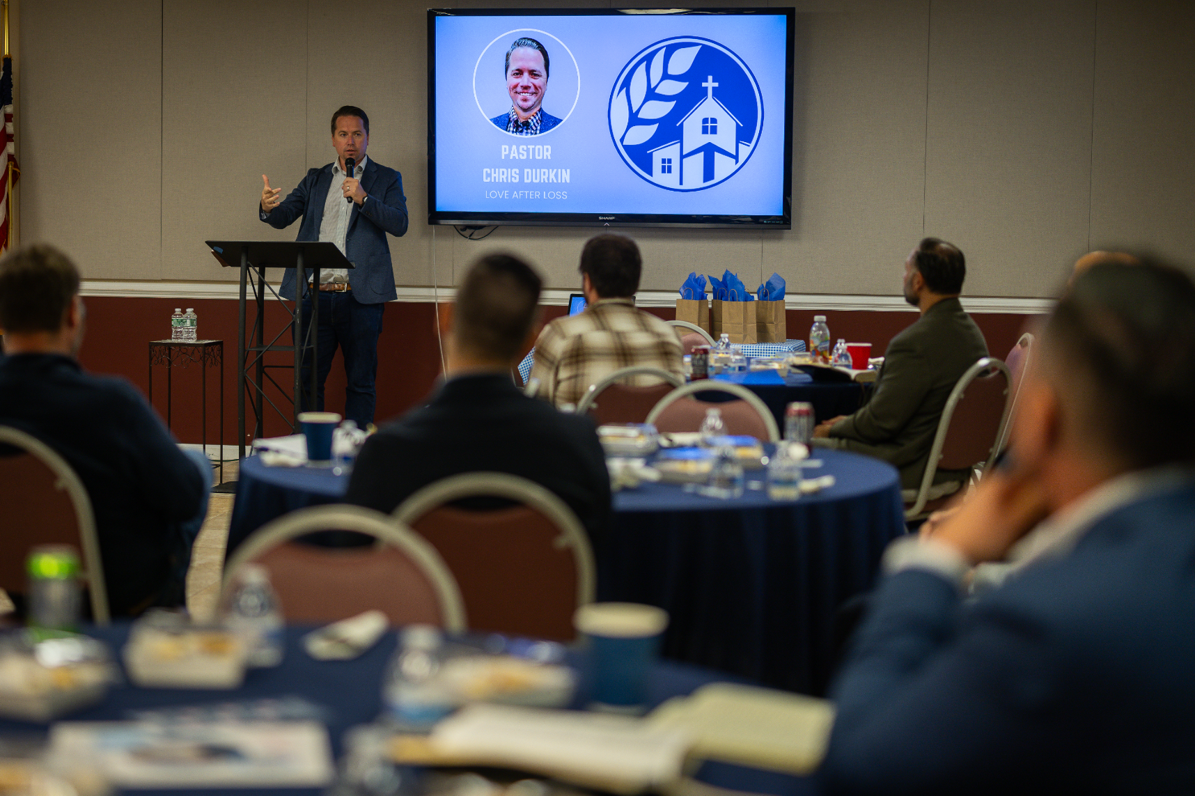 A man is giving a presentation to a group of people sitting at tables.