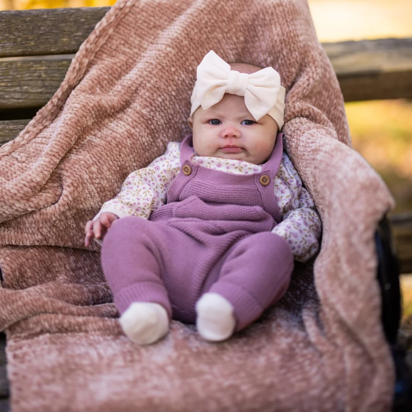 Baby in lavender overalls and bow headband, sitting on a blanket on a wooden bench.