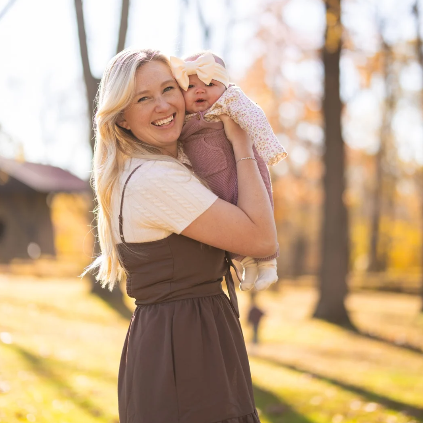 Woman smiling, holding baby. Brown dress, tan sweater. Outdoors, autumn foliage.