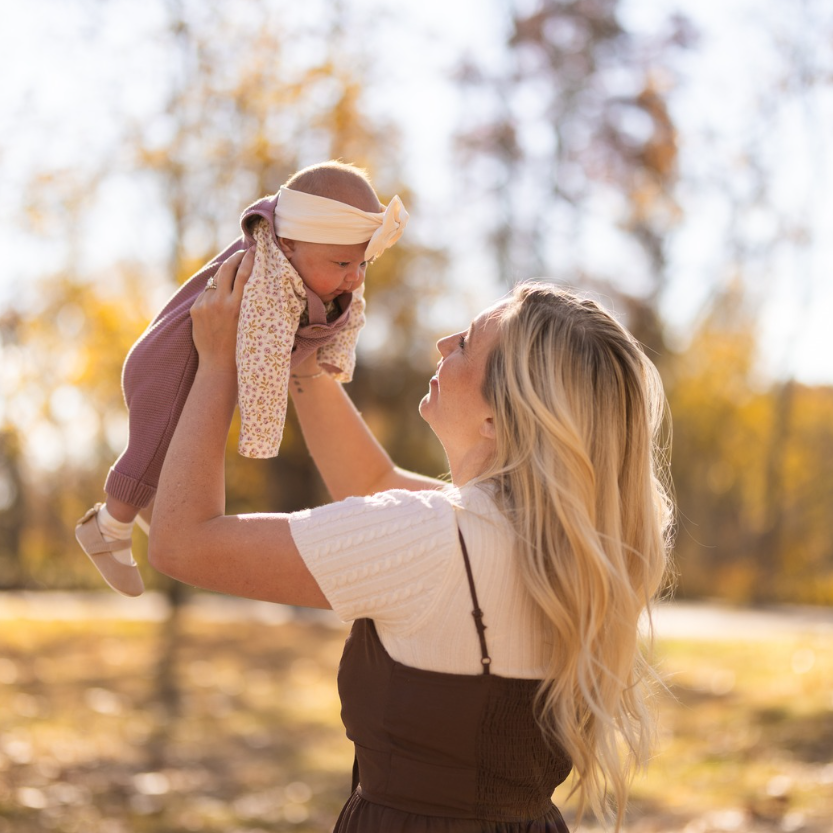 Woman holding up baby in a park with autumn foliage; baby wearing floral romper and headband.