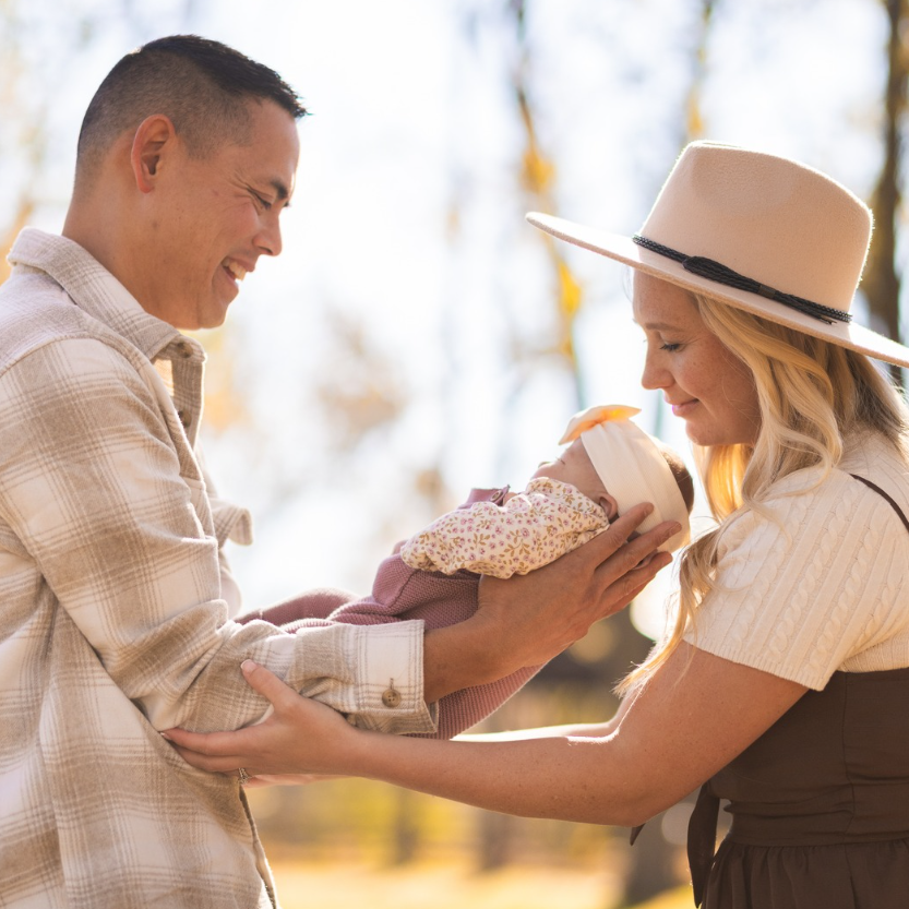 Parents holding a baby. Man smiles, woman wears a hat, set outdoors with fall foliage.